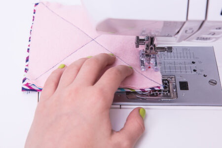 person holding a square of pink fabric under a sewing machine needle